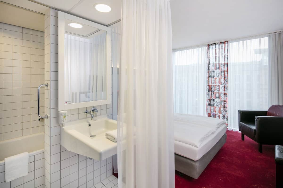 A hotel room with a bathroom, a bed, a chair, and a window with curtains.