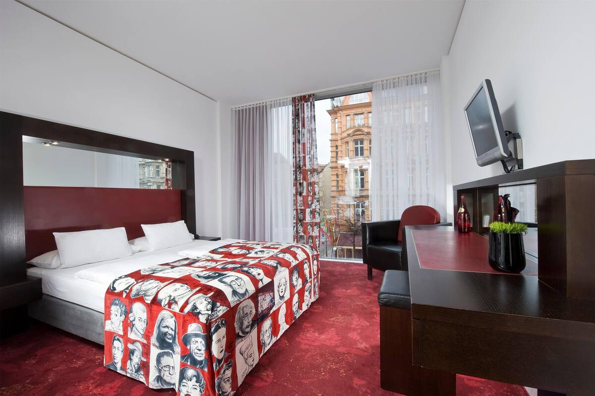 A hotel room with a large bed, a desk with a TV, and a view of buildings through the window.