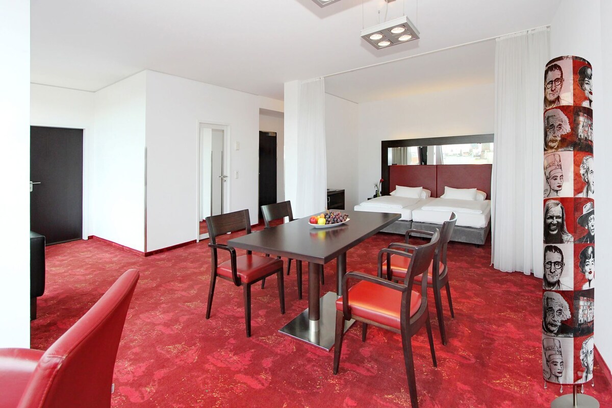 Habitación de hotel con comedor, cama con cabecera roja e impresión de arte pop en la pared.