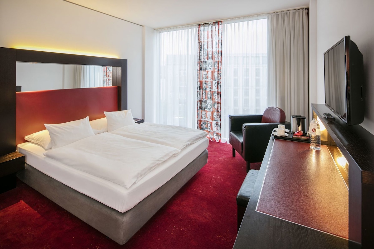 A hotel room with a large bed, a desk with a TV, a chair, and a window with curtains.