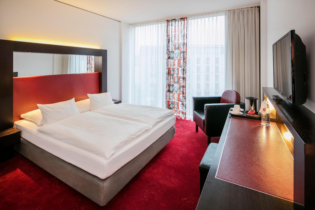 A hotel room with a large bed, a desk with a TV, a chair, and a window with curtains.