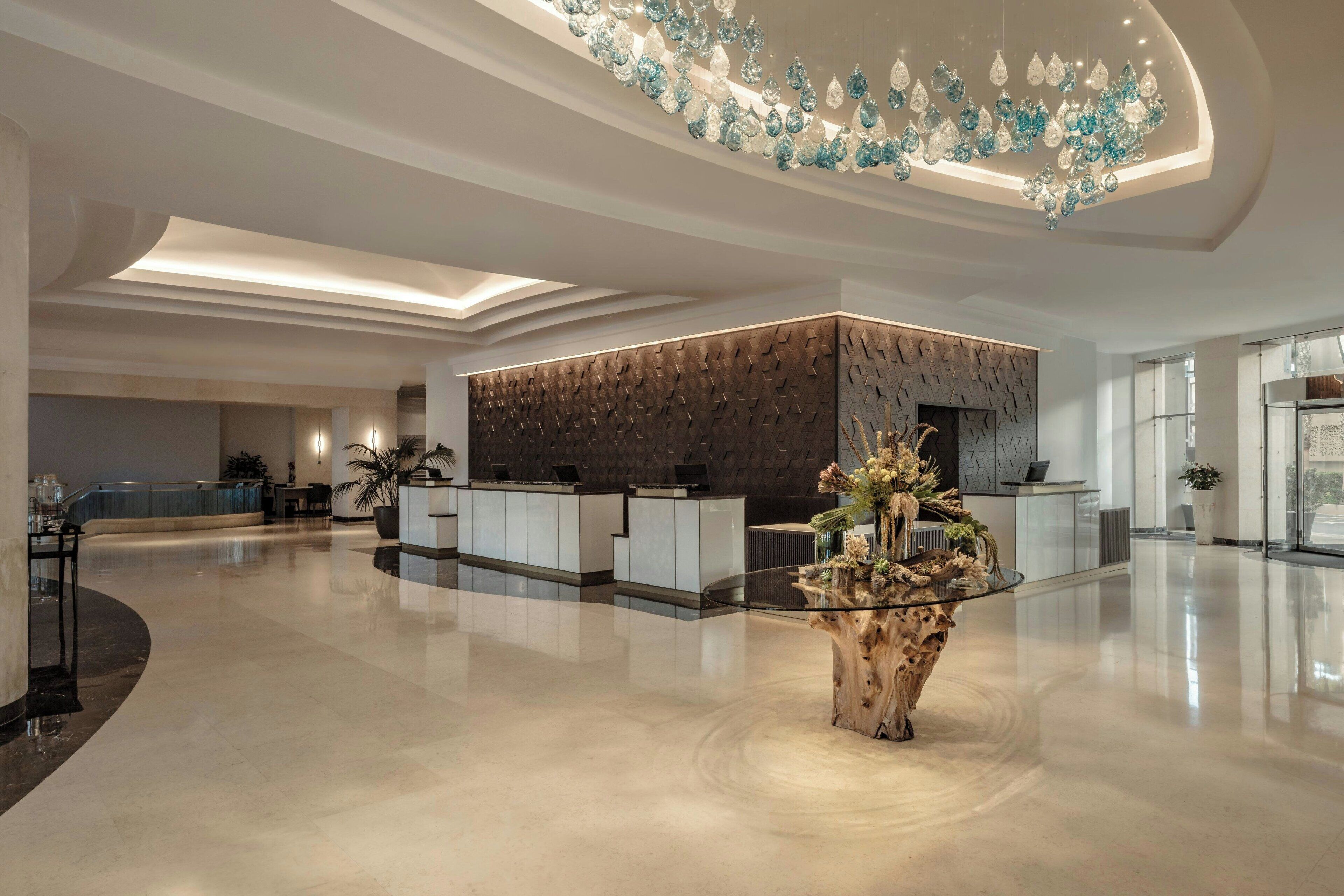 Eine moderne Hotelhalle mit einer Rezeption, einer dekorativen Deckenleuchte und einem zentral platzierten Holztisch mit Blumen.
