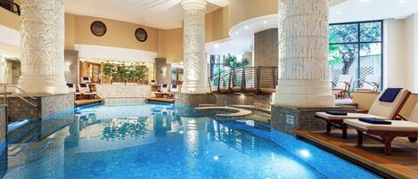 An indoor swimming pool with ornate columns, lounge chairs, and a view of the outdoors.