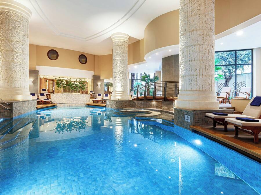 Una piscina interior con columnas ornamentales, sillones de descanso y vistas al exterior.