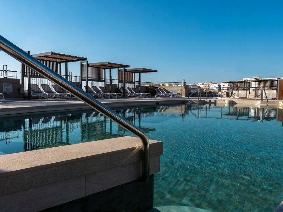 Una piscina cristalina con sillas de descanso y zonas de sombra, rodeada por una estructura de edificio moderna.