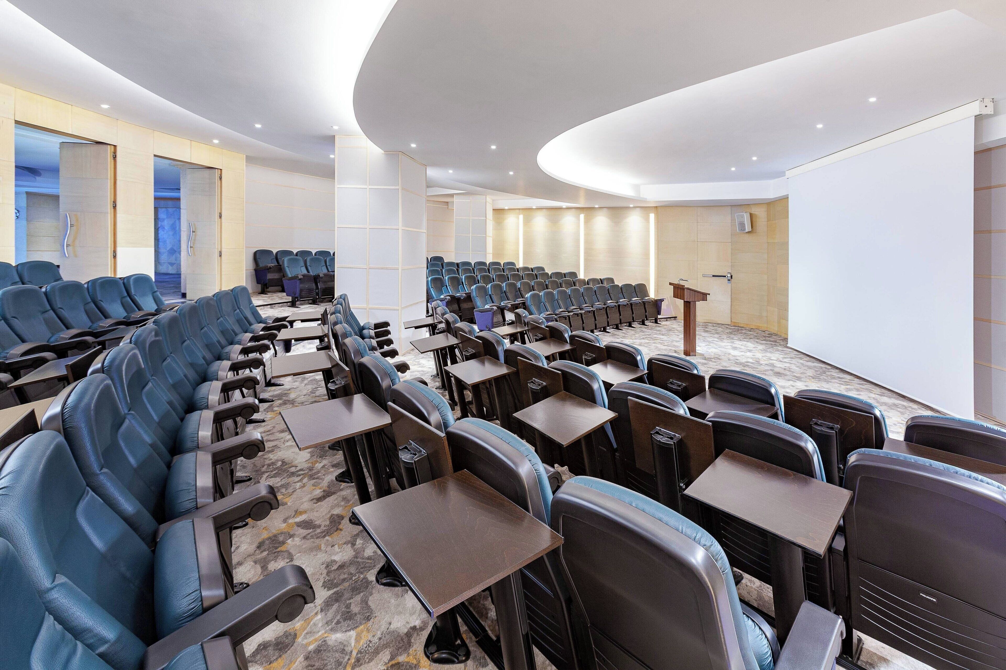 Una sala conferenze con file di sedie blu e tavoli, un podio e uno schermo bianco grande.