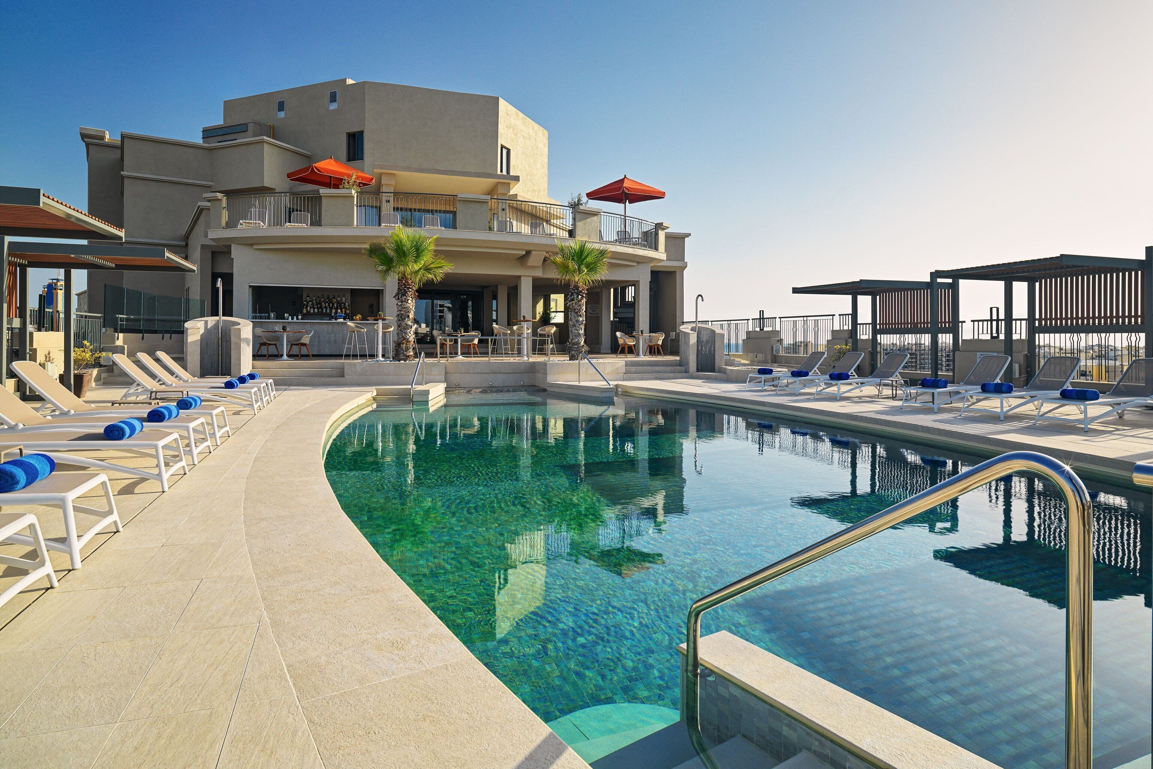 A pool area with lounge chairs and umbrellas, a bar, and a building with multiple floors and balconies.