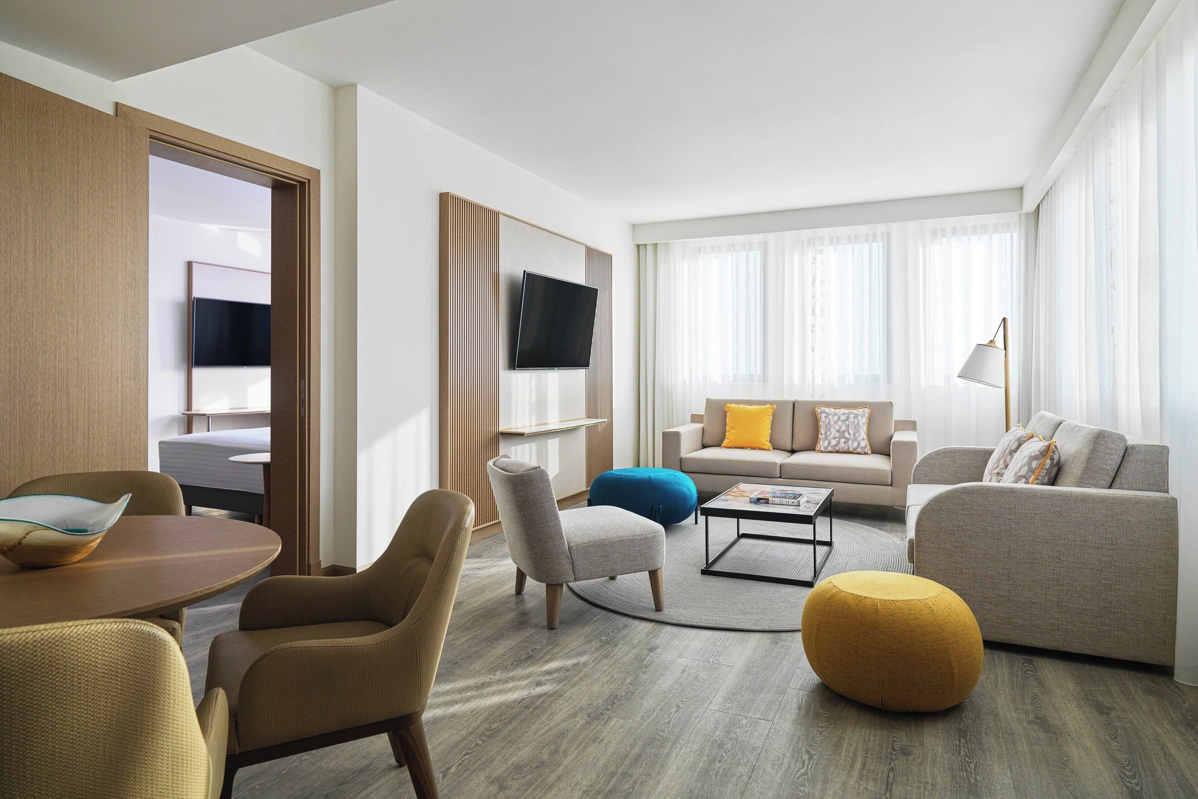 A modern hotel room with a sofa set, a dining table, a TV mounted on the wall, and a large window with sheer curtains.