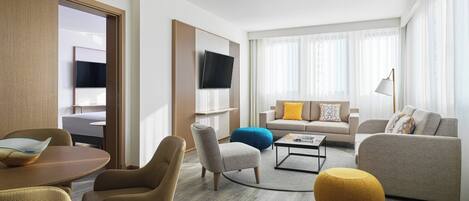 A modern hotel room with a sofa set, a dining table, a TV mounted on the wall, and a large window with sheer curtains.