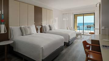 A hotel room with two beds, a desk, and a balcony with a view of the sea.