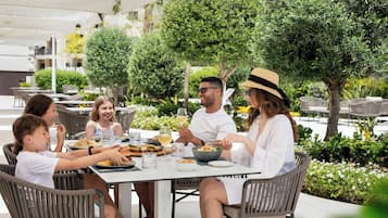 A group of people dining at an outdoor table with chairs, surrounded by greenery and trees.