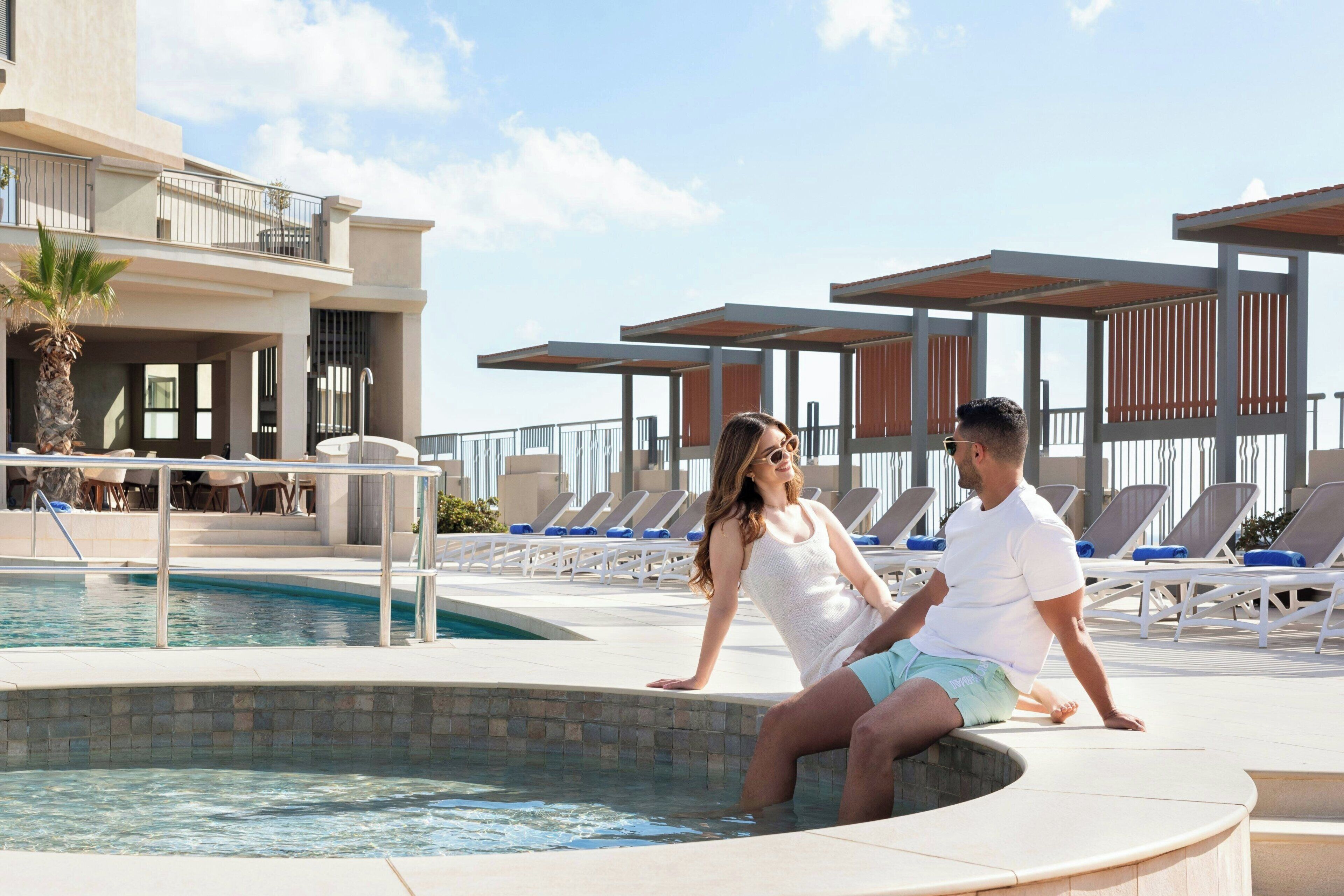 A pool area with lounge chairs, a hot tub, and a building with balconies.