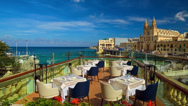 A terrace with tables and chairs overlooking a coastal area with a historic building in the background. - Malta Marriott Resort & Spa (St. Julian's)