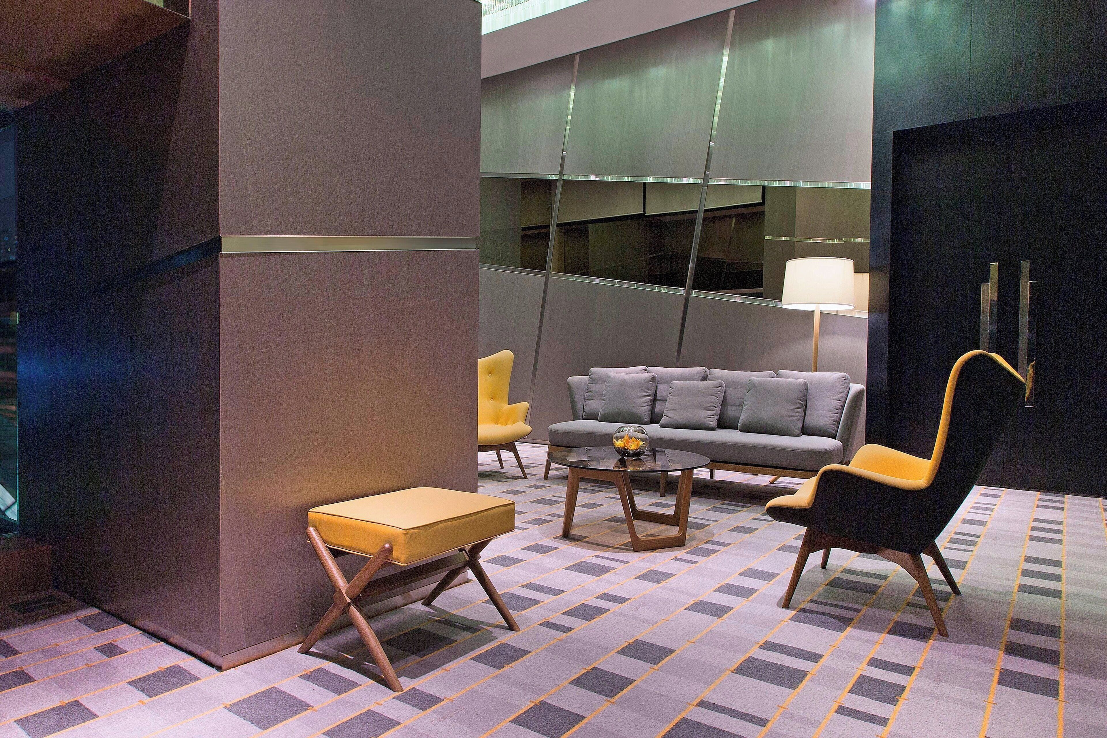 Un vestíbulo moderno con un sofá gris, un sillón amarillo y un sillón negro. Hay una mesita con una lámpara y una alfombra estampada.