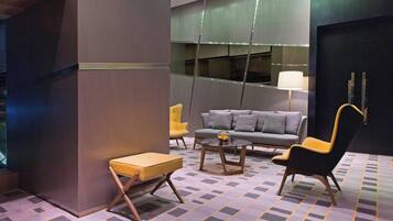 Un vestíbulo moderno con un sofá gris, un sillón amarillo y un sillón negro. Hay una mesita con una lámpara y una alfombra estampada.