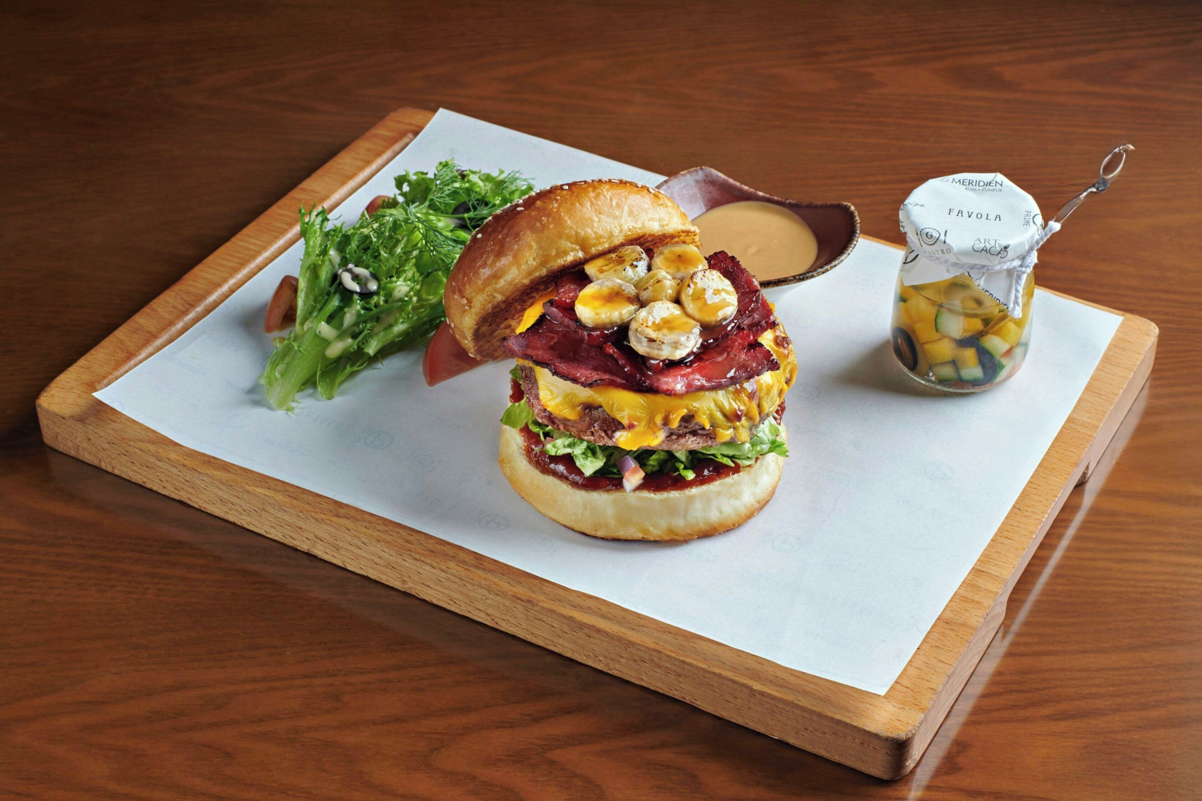 A sandwich on a wooden board with a side salad and a small jar of pickles.