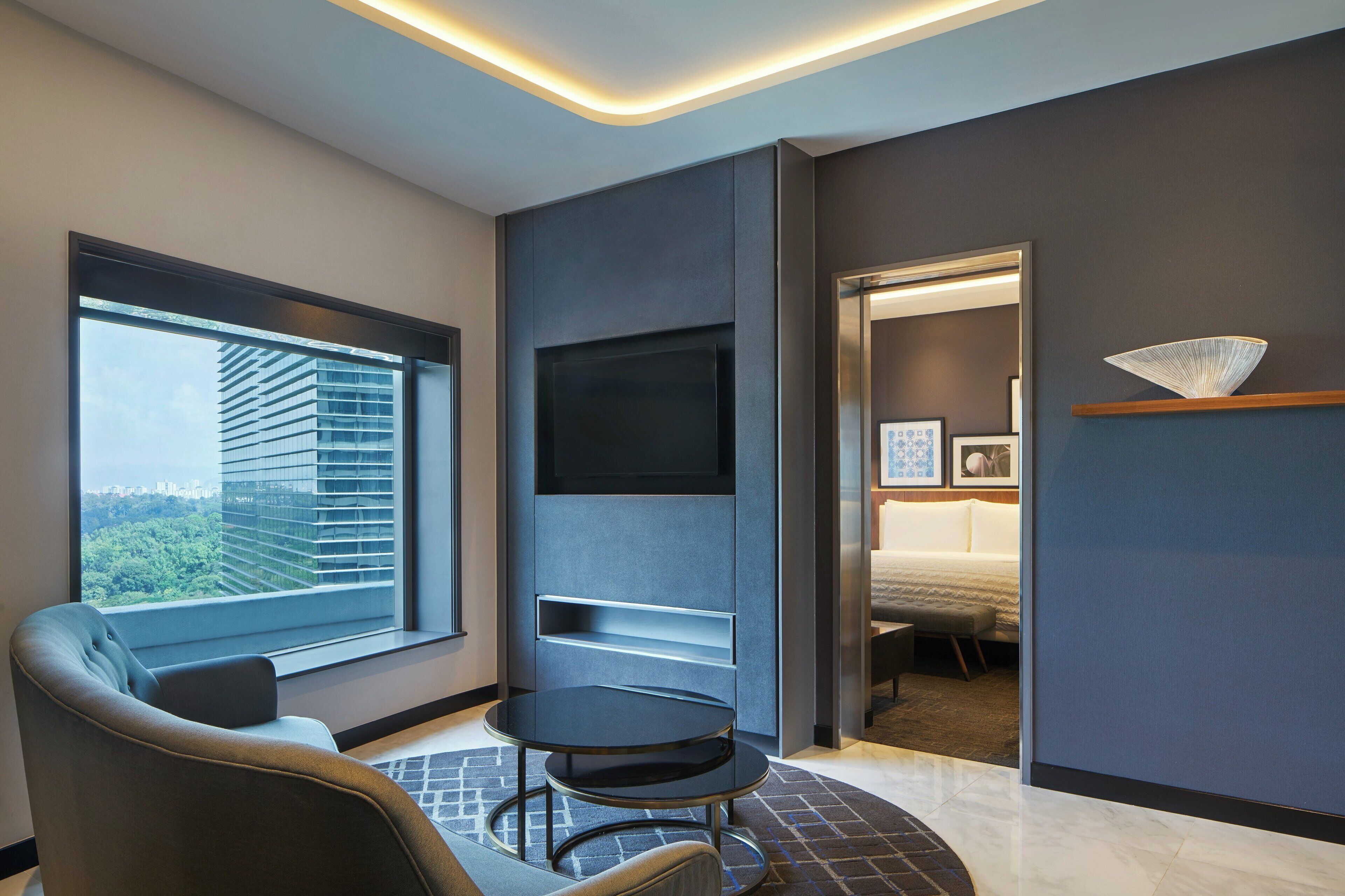 Une chambre d’hôtel moderne dotée d’une grande fenêtre, d’un téléviseur à écran plat fixé au mur, d’une table ronde et d’un coin salon confortable.
