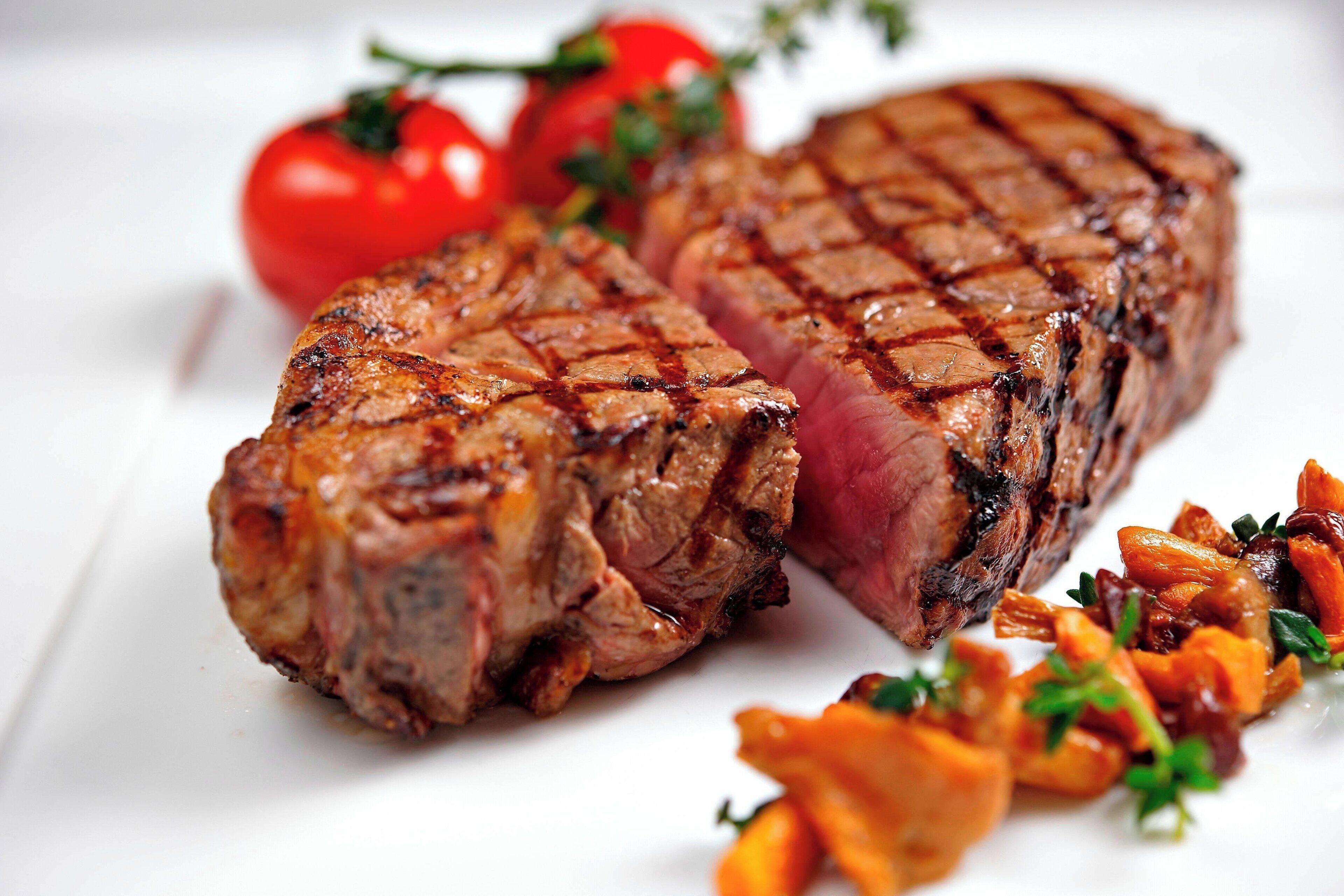 A grilled steak with a charred exterior, accompanied by grilled vegetables and fresh tomatoes.
