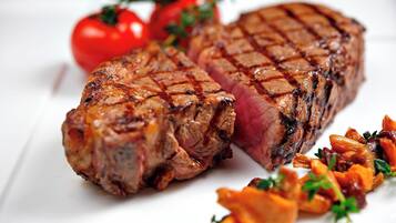 Un bistec a la parrilla con una capa exterior quemada, acompañado de verduras asadas y tomates frescos.