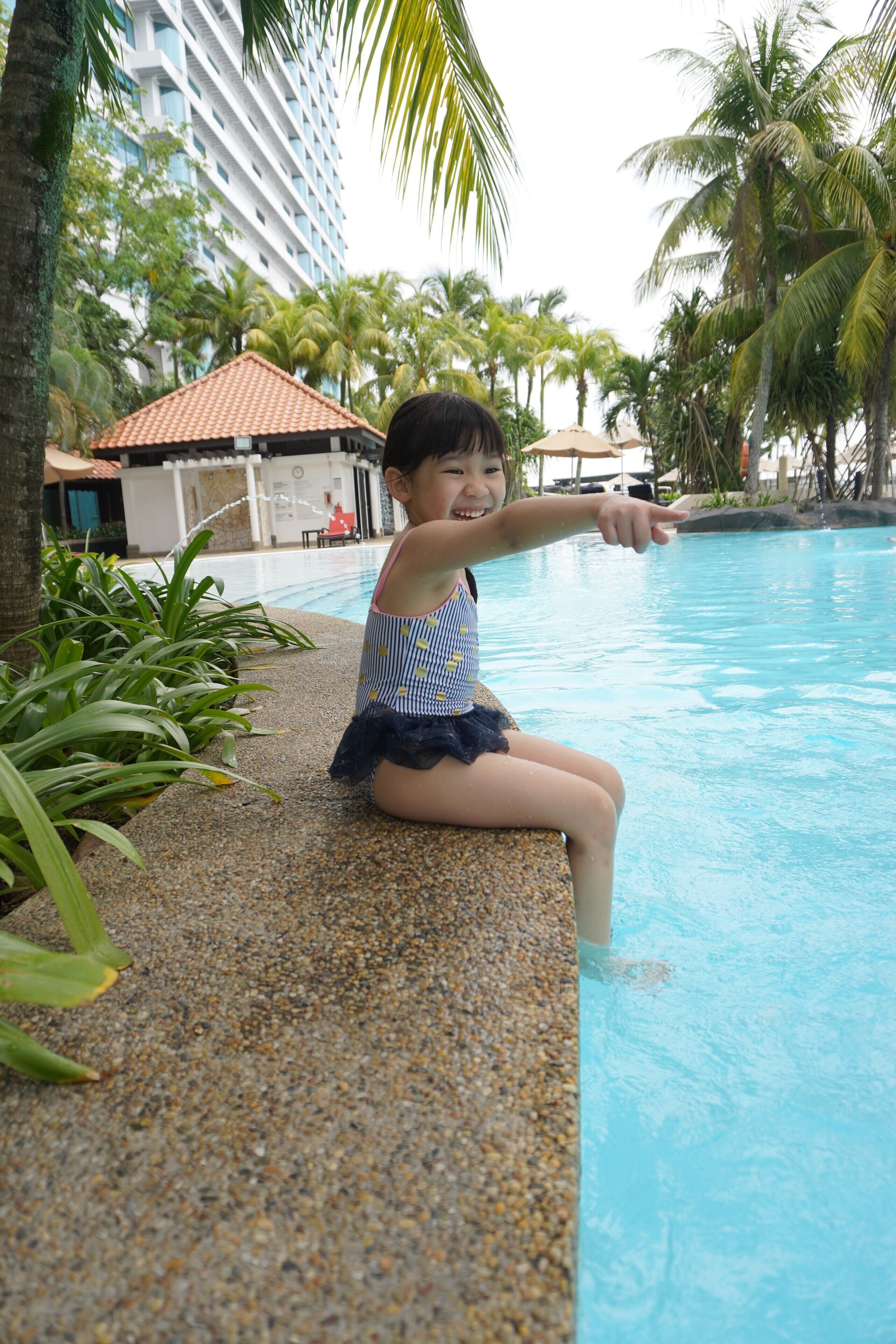 A child sitting by a pool with palm trees and a building in the background.
