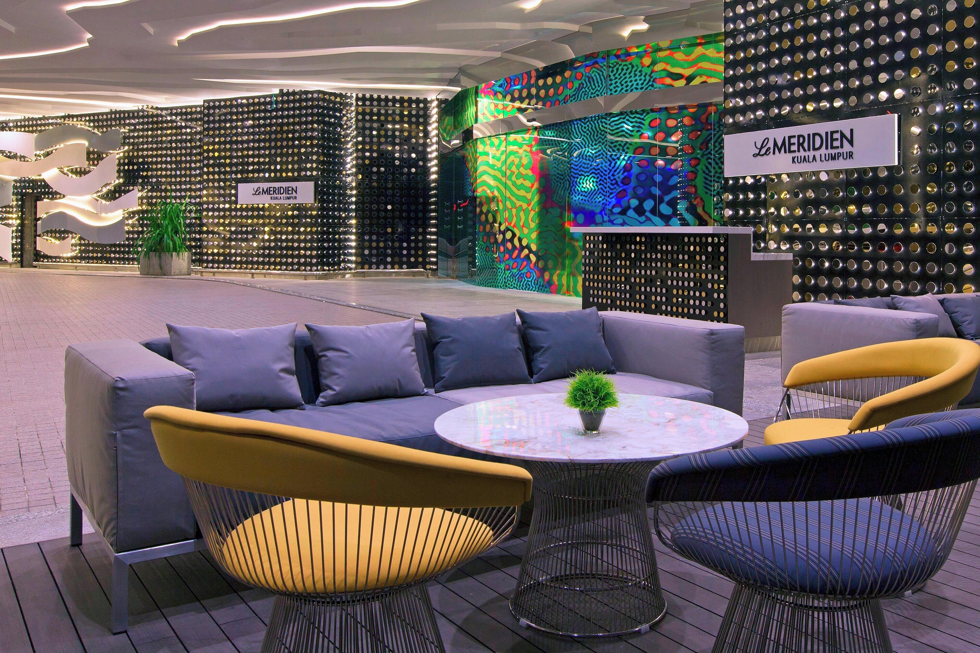 Ein moderner Lounge-Bereich mit einer Couch, Sesseln und einem kleinen Tisch, mit einer Wanddekoration und einem Schild mit der Aufschrift 'Le Meridien Kuala Lumpur'.