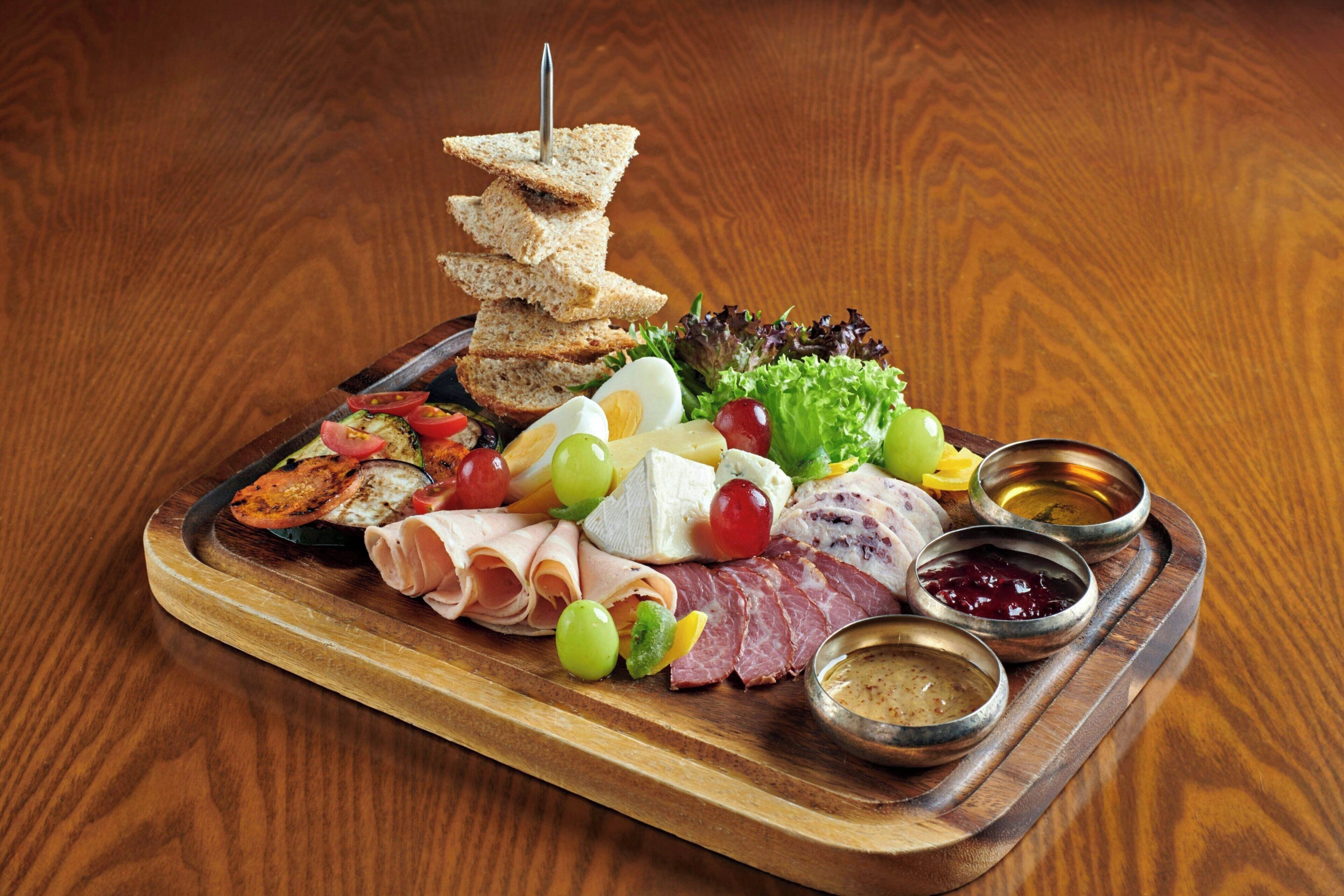 A wooden board with a variety of food items including meats, cheeses, and vegetables, accompanied by small bowls of condiments.