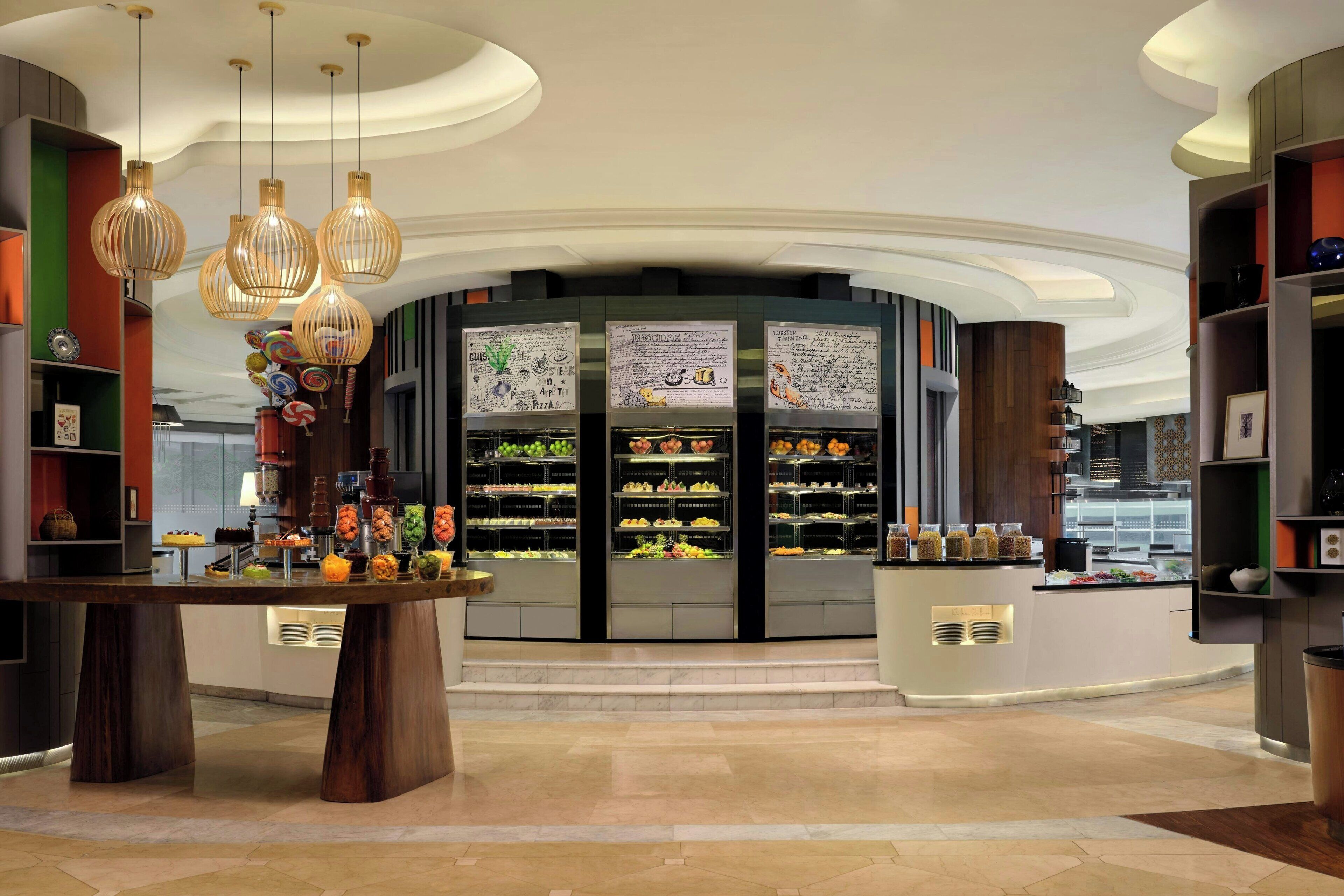 A modern hotel lobby with a central dessert display, wooden furniture, and a curved ceiling design.
