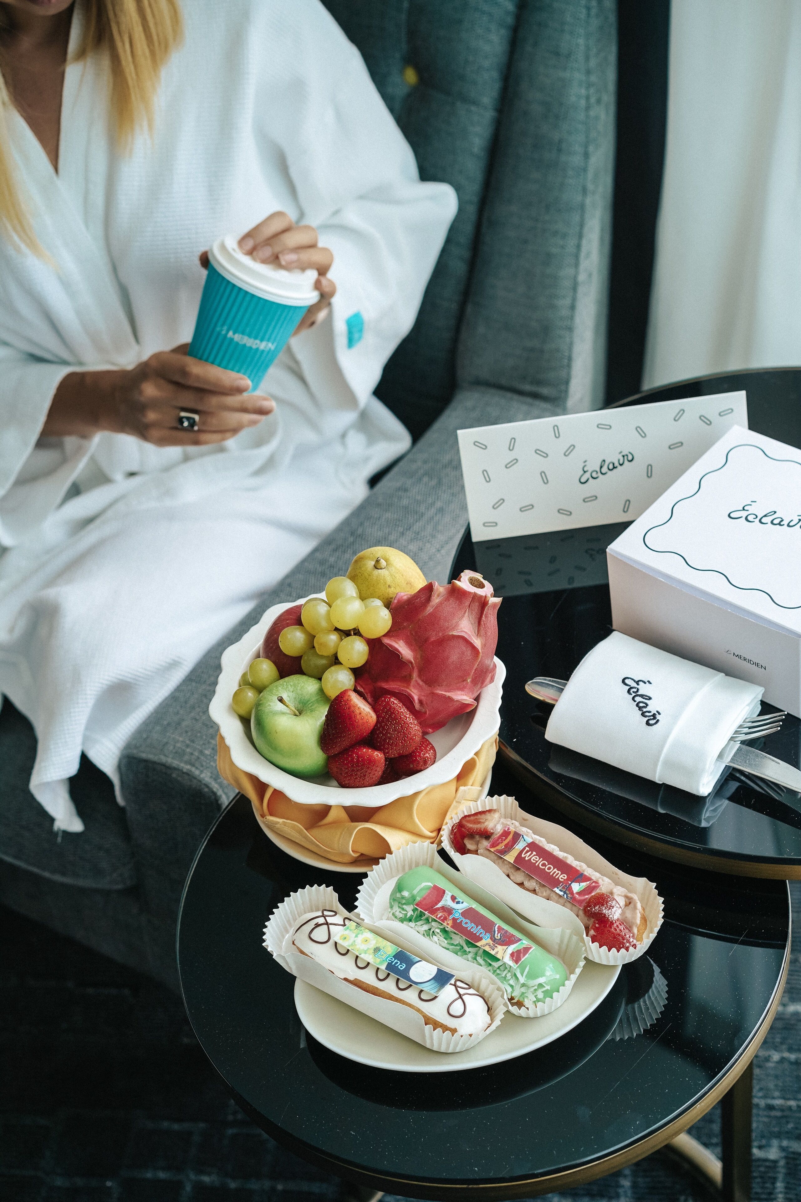 A person sitting on a couch with a coffee cup, a bowl of fruit, a plate of pastries, and a card with the name "Éclair".