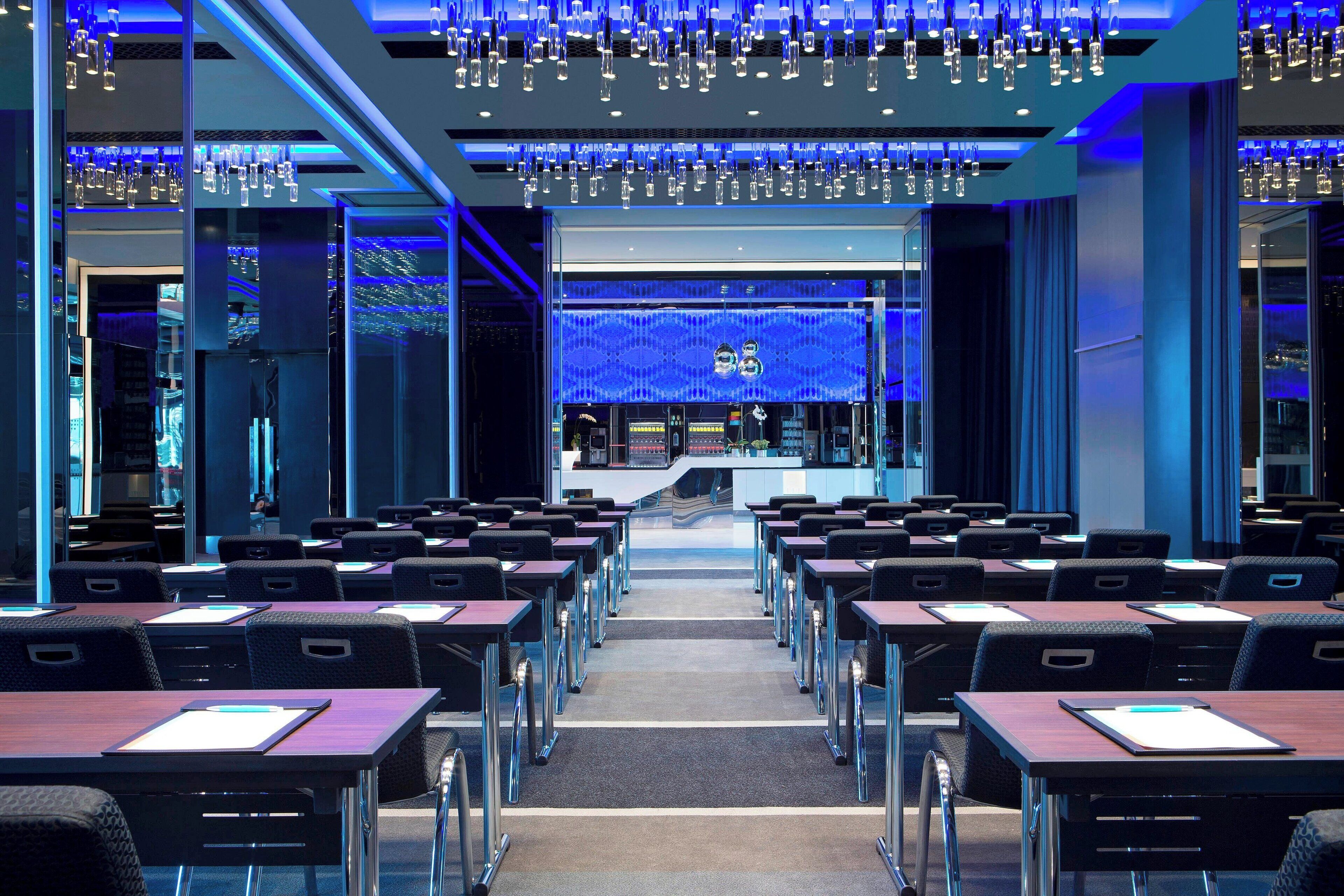 A modern conference room with rows of tables and chairs, a reception desk, and a blue-lit ceiling.