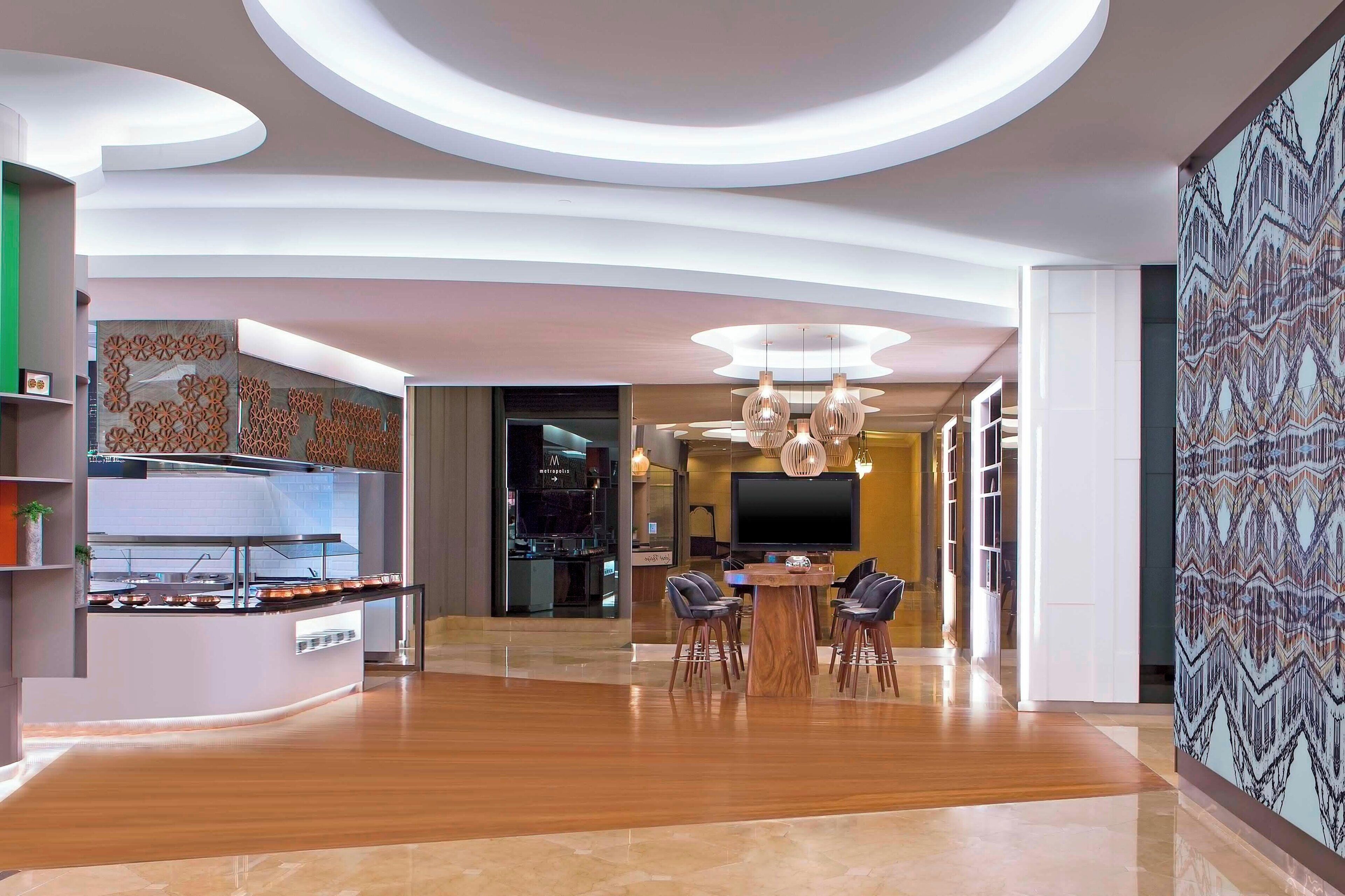Um hall de hotel moderno com área de refeições central, cozinha e uma grande tela de televisão.