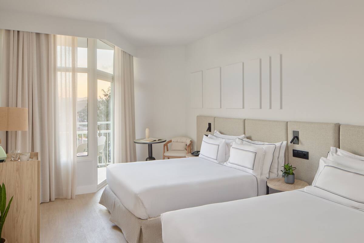 Ein Hotelzimmer mit zwei Betten, einem Schreibtisch, einem Sessel, einem kleinen Tisch und einer Topfpflanze.
