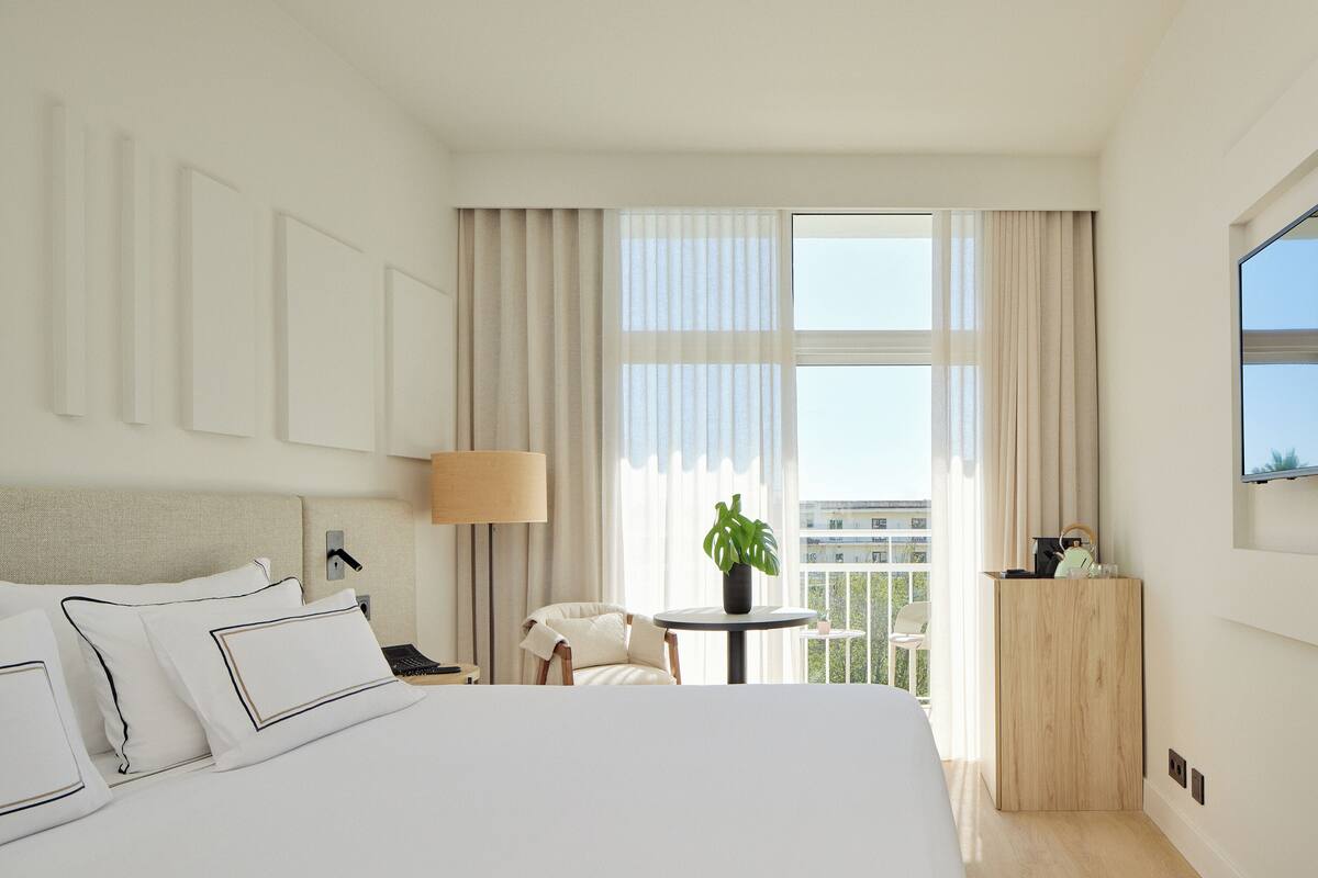 Ein Hotelzimmer mit einem großen Bett, einem kleinen runden Tisch mit einer Pflanze, einem Sessel und Blick auf die Stadt durch große Fenster.