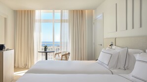 A hotel room with a large bed, a desk with a chair, a small table with a plant, and a view of the sea through large windows. - Melia Lloret de Mar (Lloret de Mar)