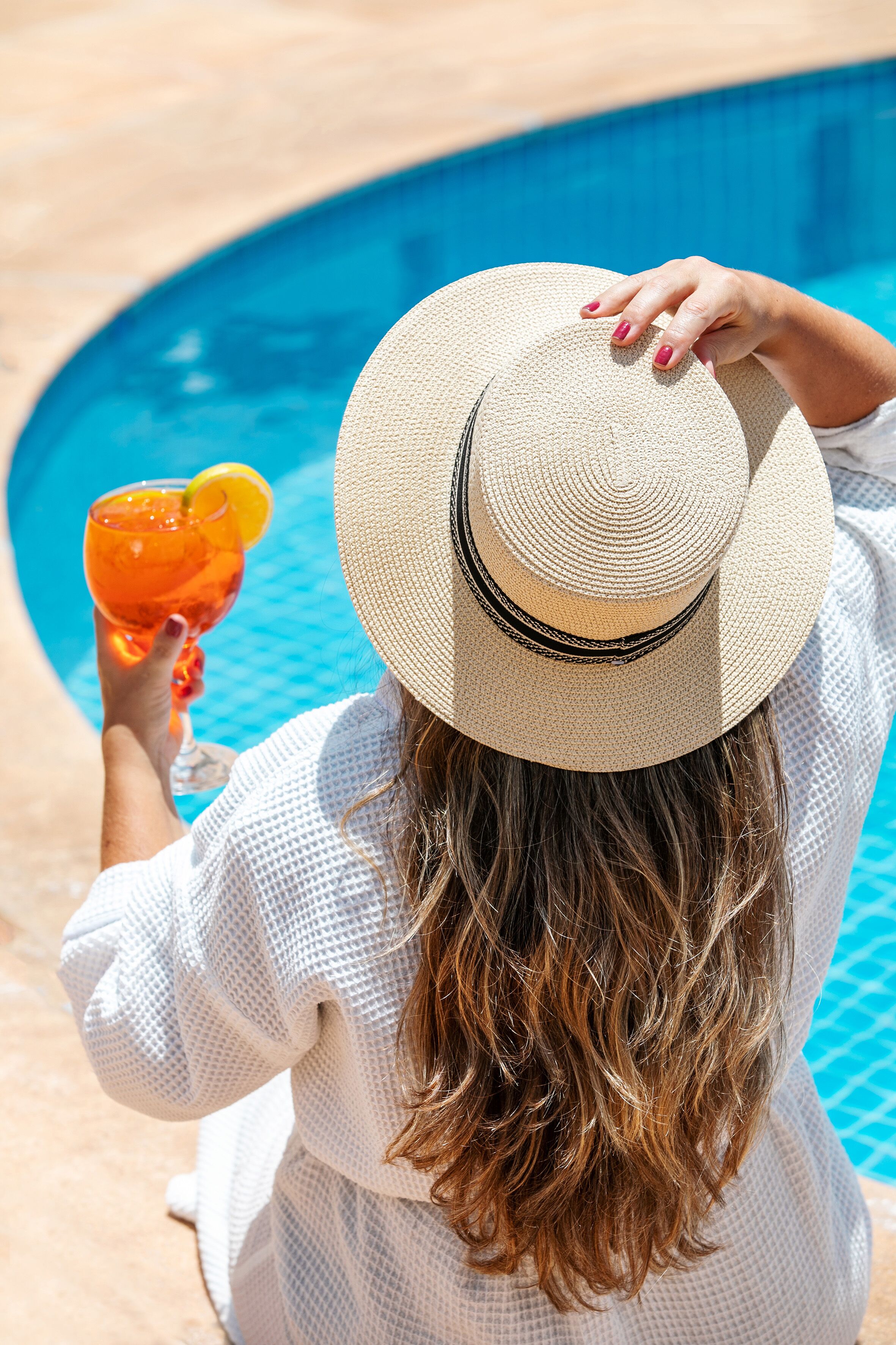 A person sitting by a pool with a drink, wearing a hat and a white outfit.