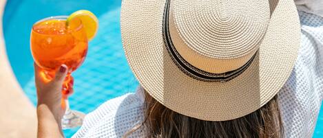 A person sitting by a pool with a drink, wearing a hat and a white outfit.