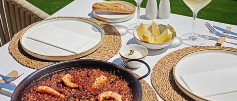 A table set for a meal with a large pan of seafood paella, plates, bread, and glasses of white wine.