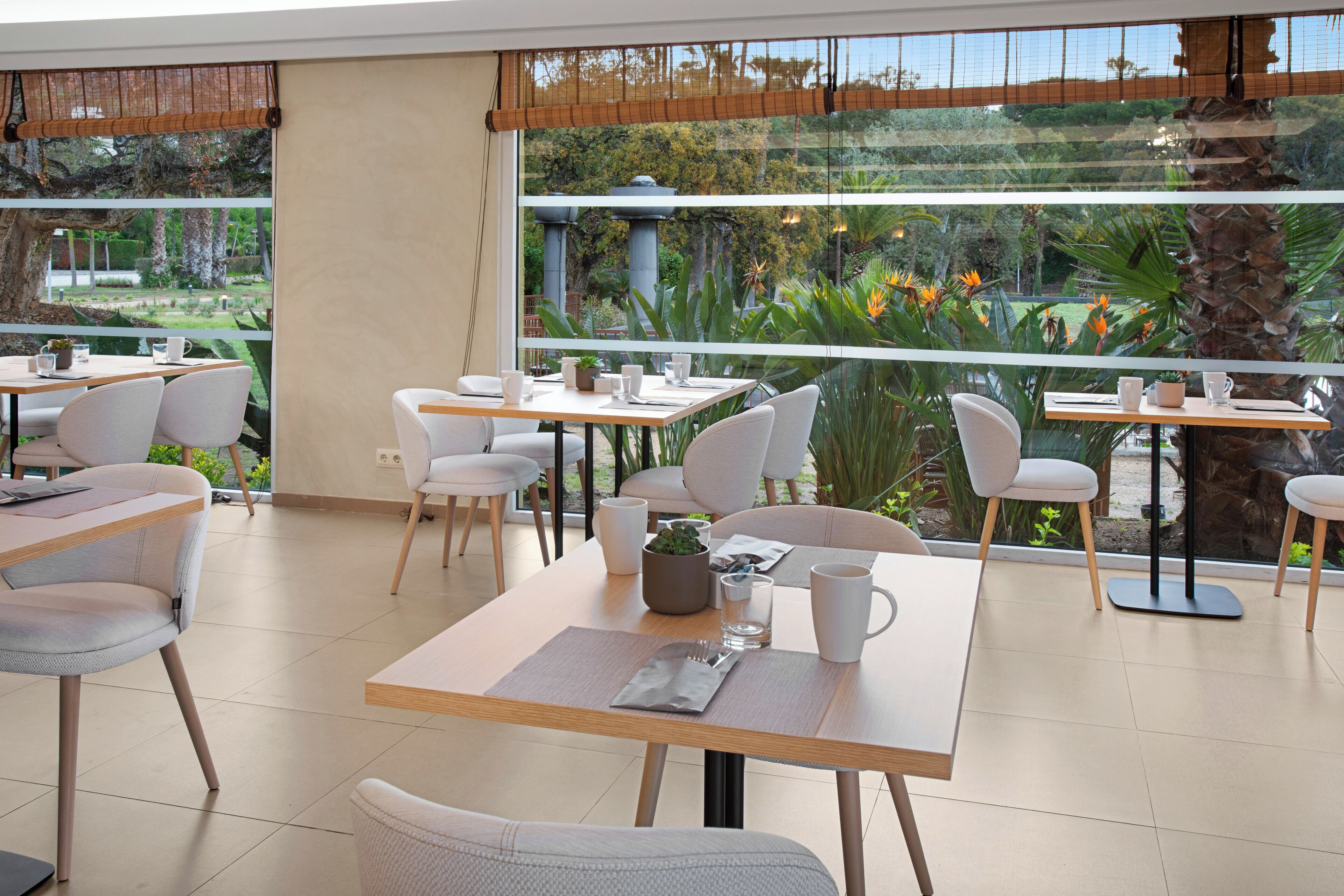 A restaurant with multiple tables and chairs, large windows with a view of greenery and a palm tree, and a modern interior design.