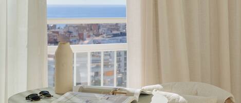 A balcony with a view of the city and the sea, a round table with a vase and sunglasses, and a cushioned chair.