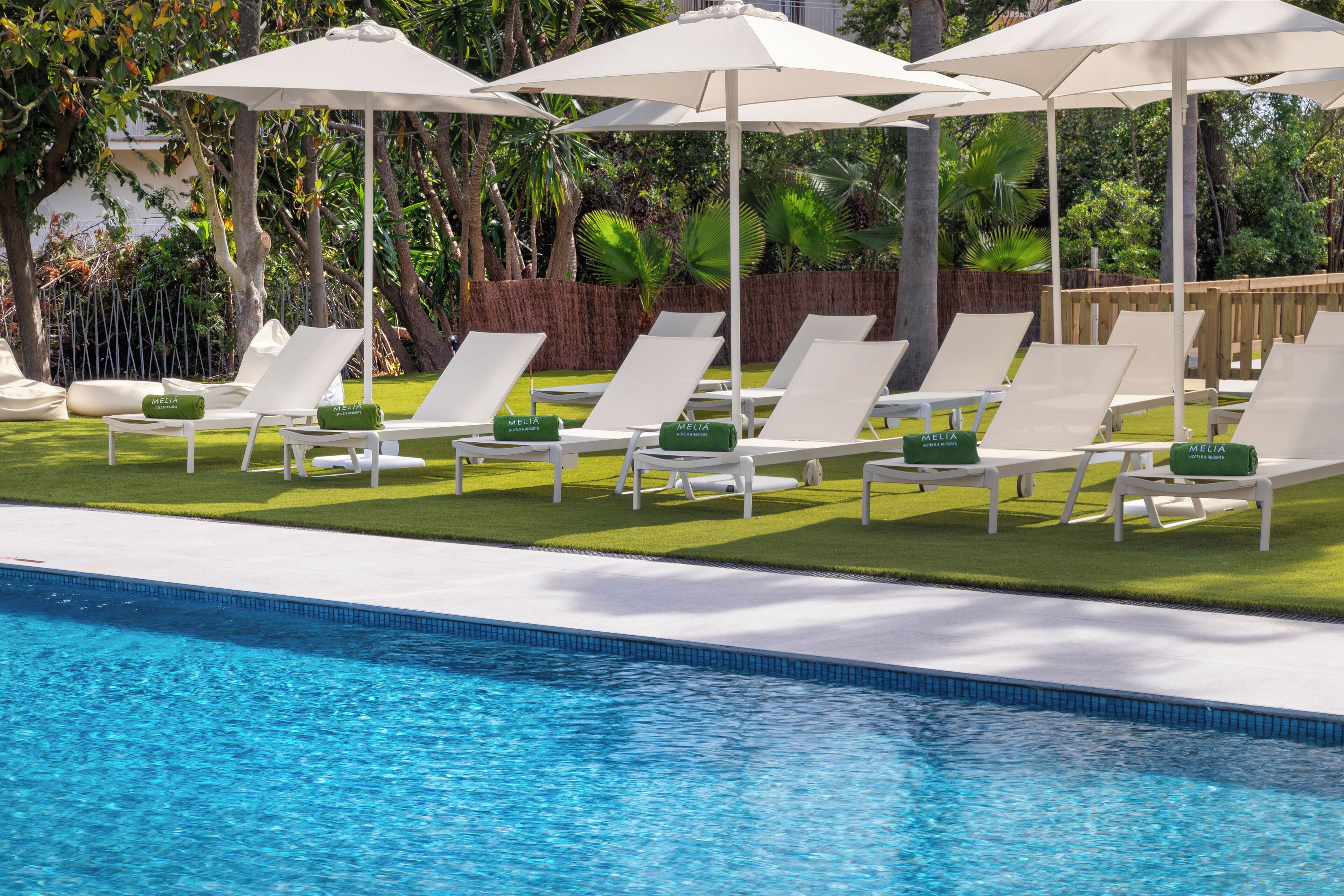 A poolside area with white lounge chairs and umbrellas, surrounded by greenery.