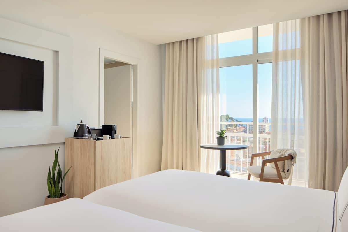 Ein Hotelzimmer mit einem großen Bett, einem kleinen Tisch mit einer Pflanze, einem Sessel, einem an der Wand montierten Fernseher und einem Fenster mit Vorhängen.