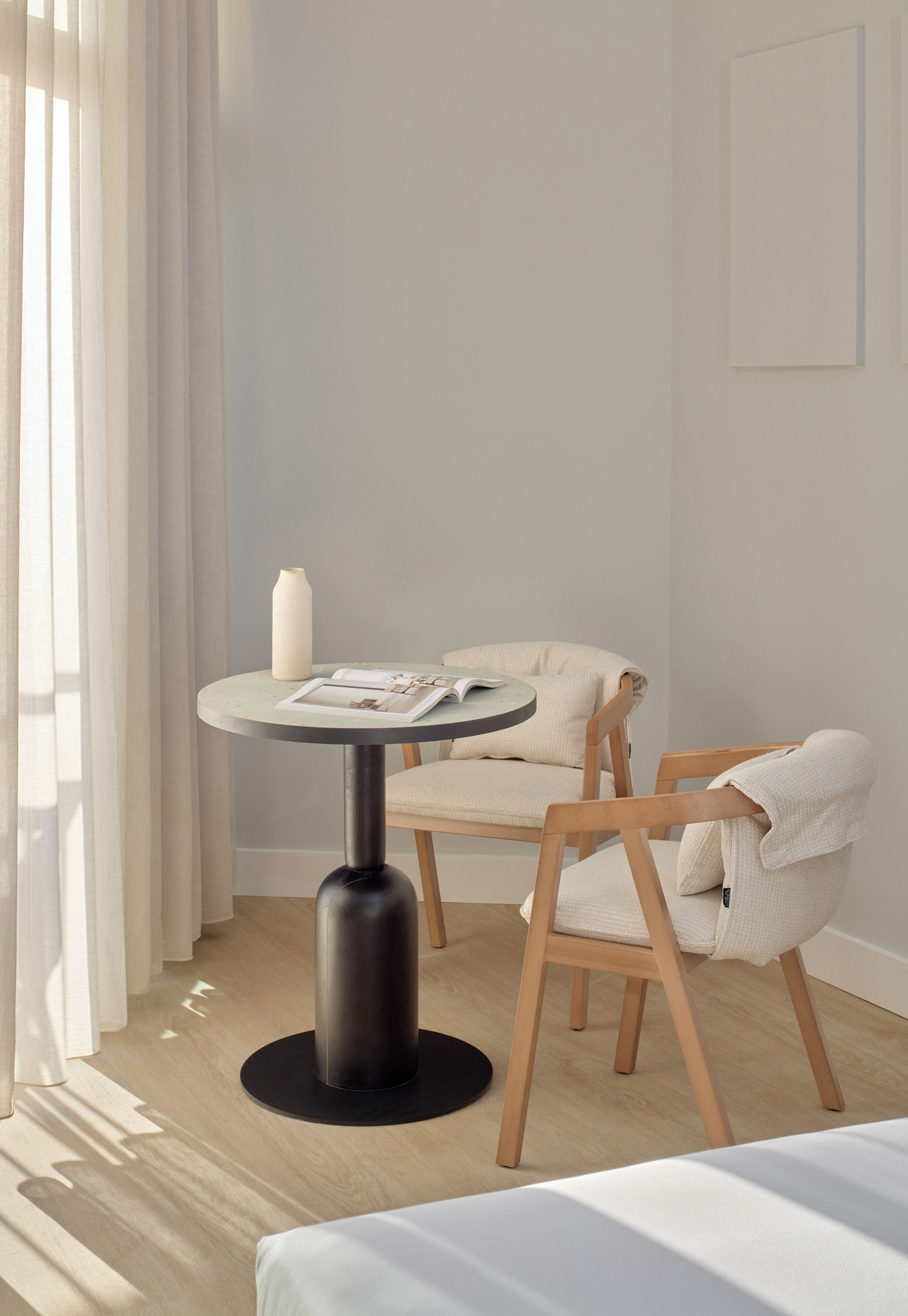 A minimalist room with a round table, two wooden chairs, a white vase, and a framed picture on the wall.