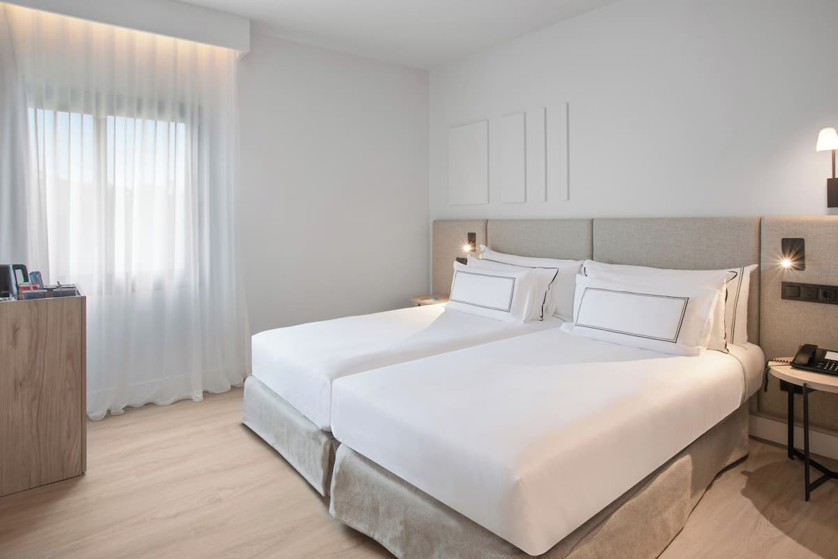 Ein Hotelzimmer mit zwei Betten, einem Nachttisch mit Lampe, einer Holzkommode und einem Fenster mit durchsichtigen Vorhängen.