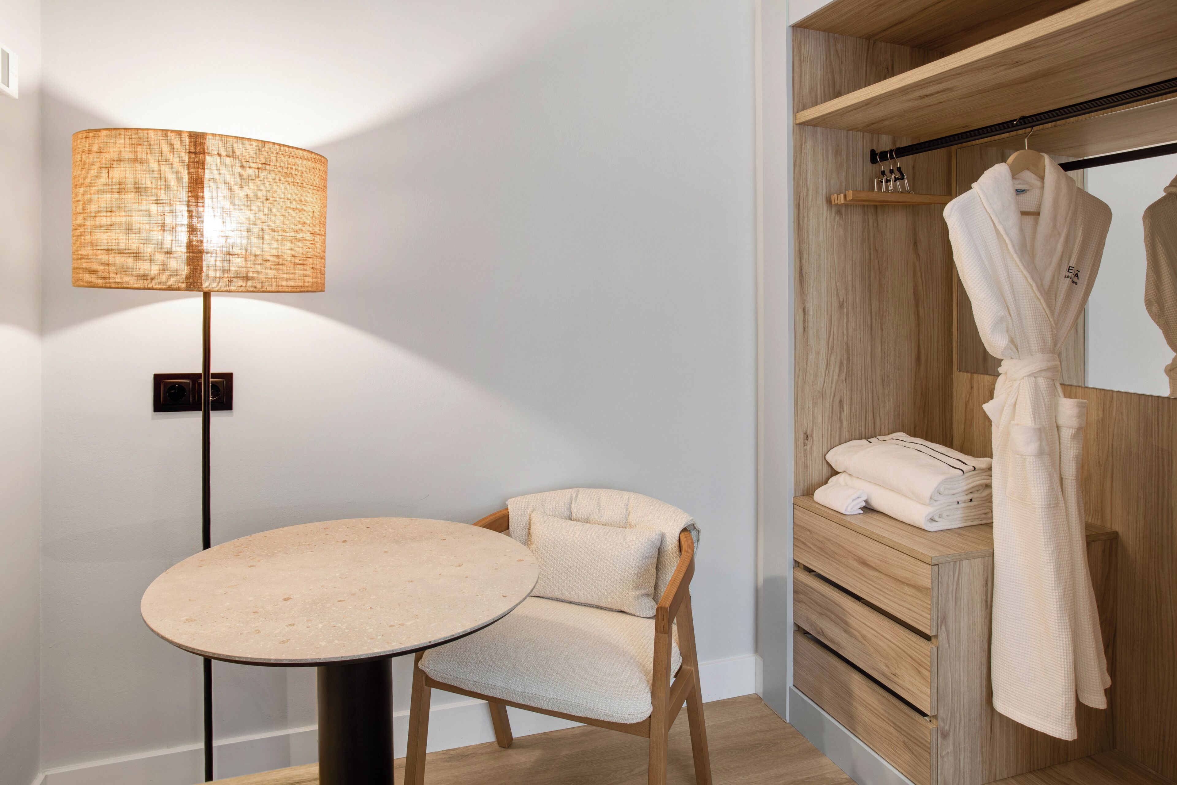 A modern bathroom with a round table, a chair, a standing lamp, and a wooden vanity with a robe hanging.