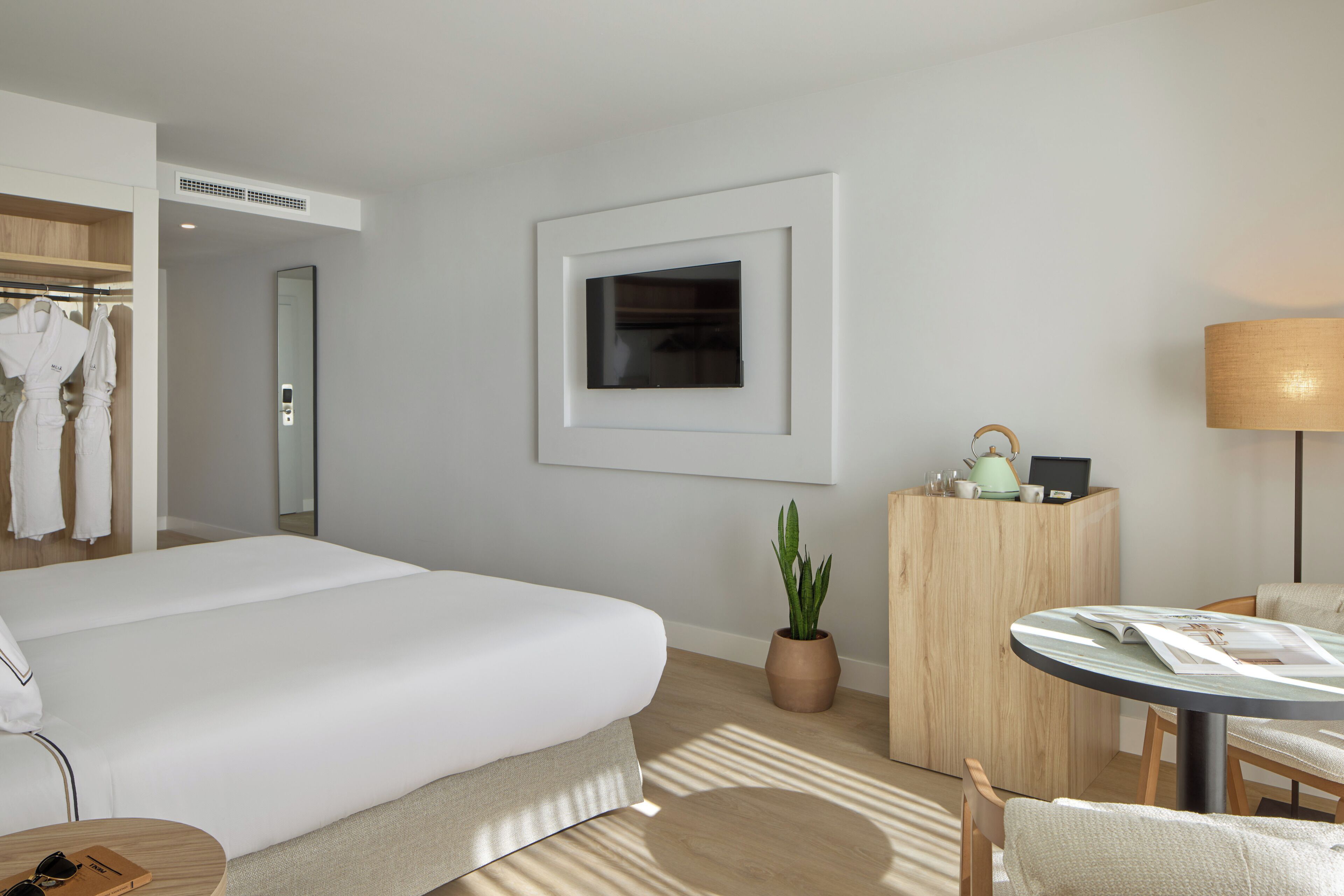 Habitación de hotel moderna con una cama grande, una mesita de noche con lámpara, un escritorio pequeño con una computadora portátil, una planta en maceta y un armario con batas.