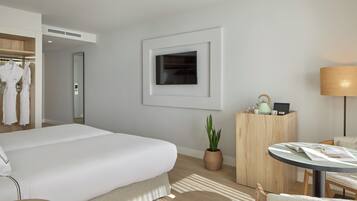 Habitación de hotel moderna con una cama grande, una mesita de noche con lámpara, un escritorio pequeño con una computadora portátil, una planta en maceta y un armario con batas.