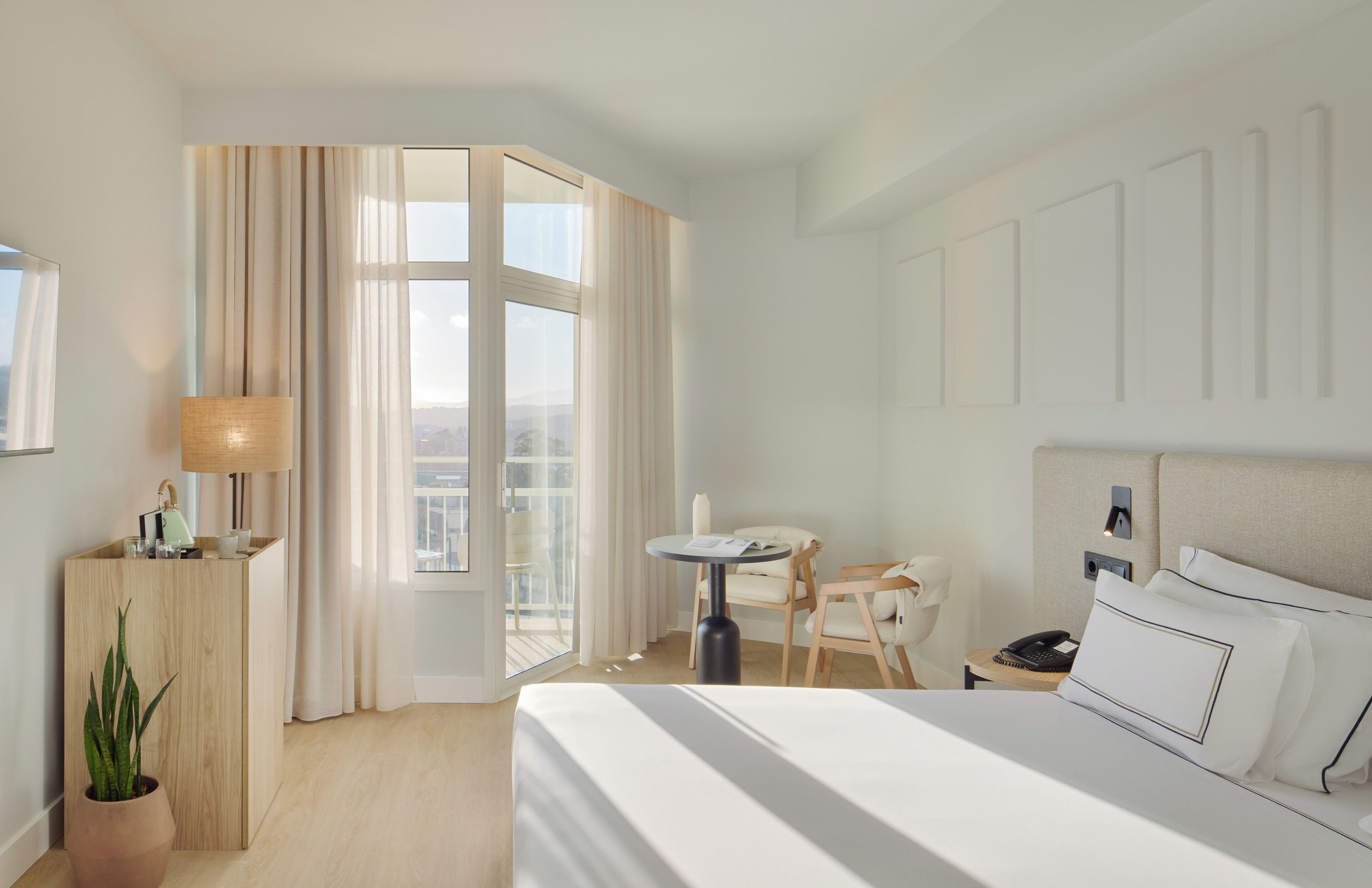 A hotel room with a large bed, a desk with a lamp, a chair, a small table, and a potted plant.