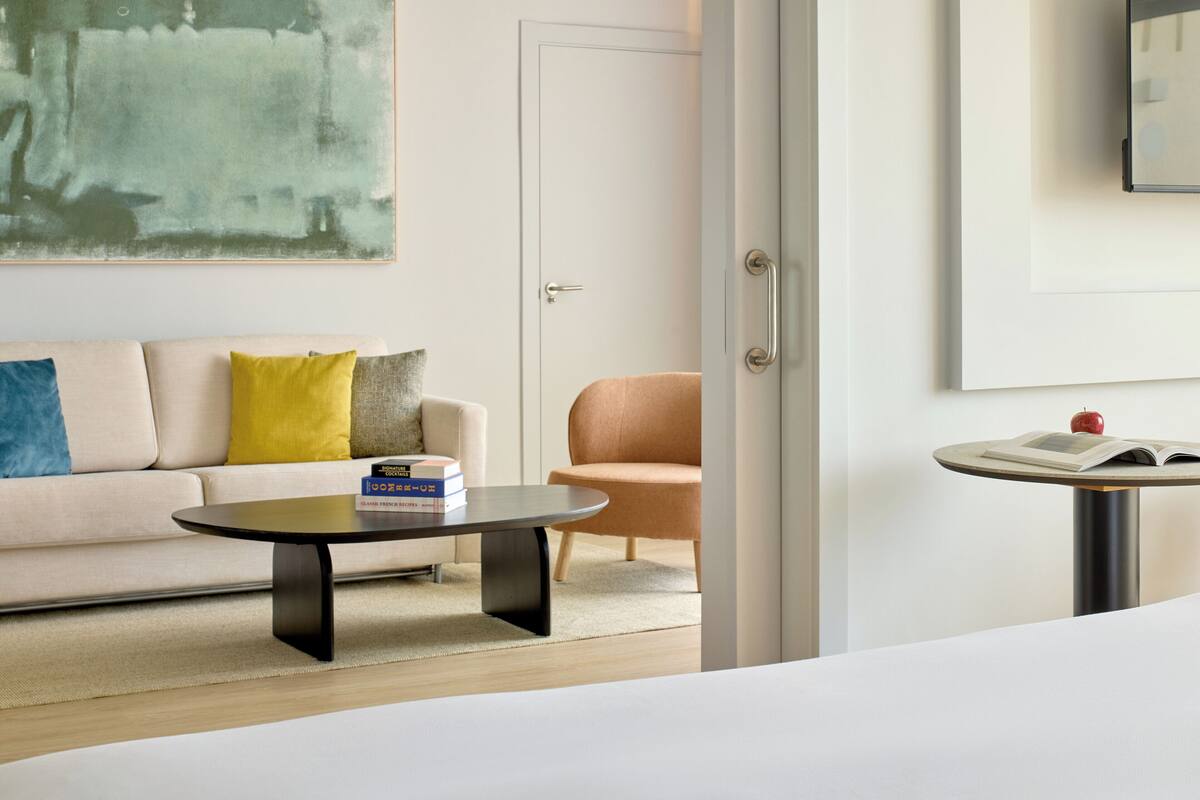 Ein modernes Hotelzimmer mit einer Couch, einem runden Couchtisch, einem Sessel und einem kleinen Tisch mit einem Buch und einem Apfel.