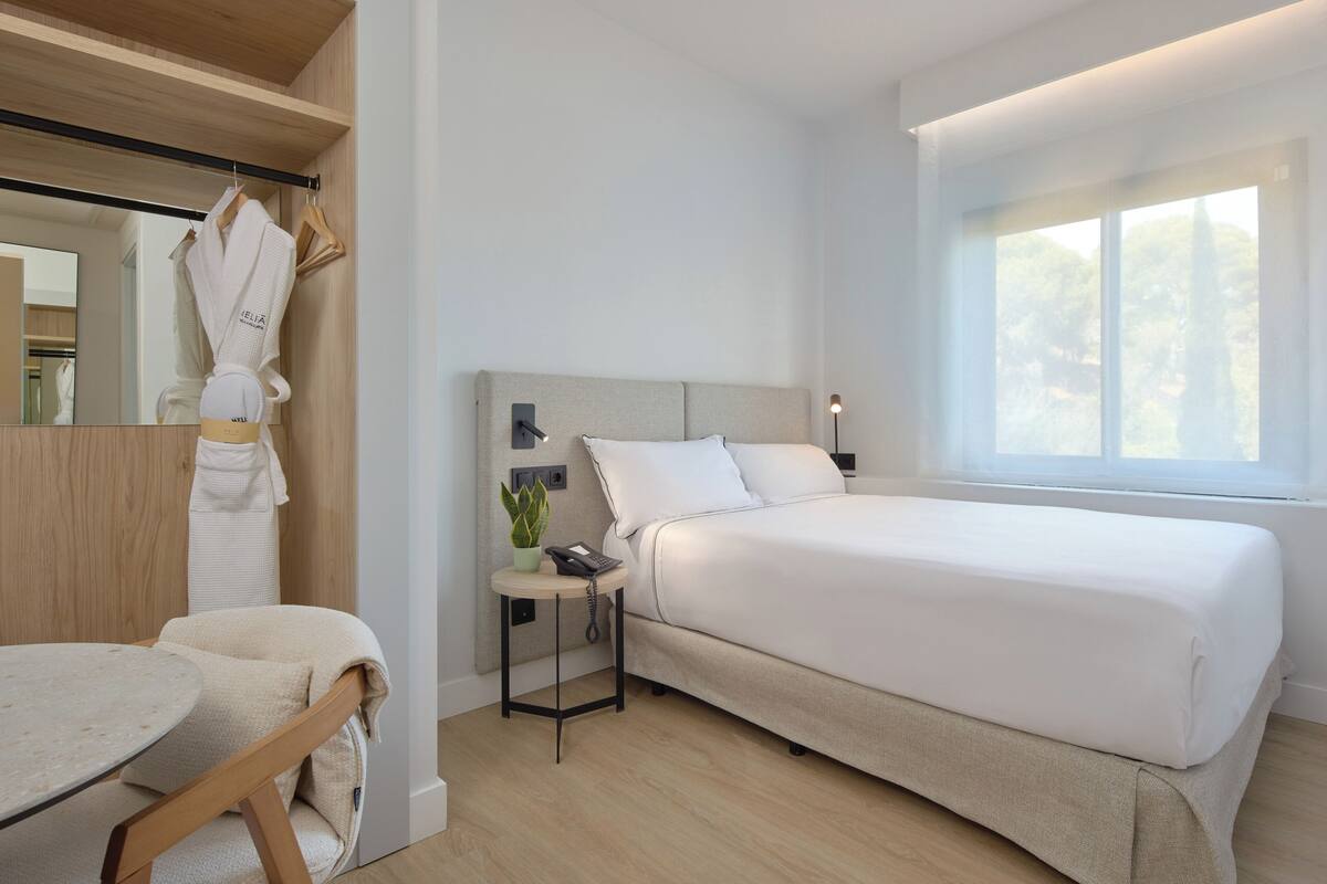 Ein modernes Hotelzimmer mit einem großen Bett, einem Nachttisch mit Lampe, einem kleinen Tisch mit Stuhl und einem Fenster mit Blick auf Bäume.