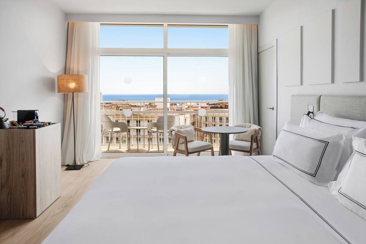 Ein Hotelzimmer mit einem großen Bett, einem Schreibtisch, einer Lampe und Blick auf Stadt und Meer durch große Fenster.