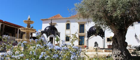 A white building with a tiled roof, a fountain, and a large tree in the foreground.